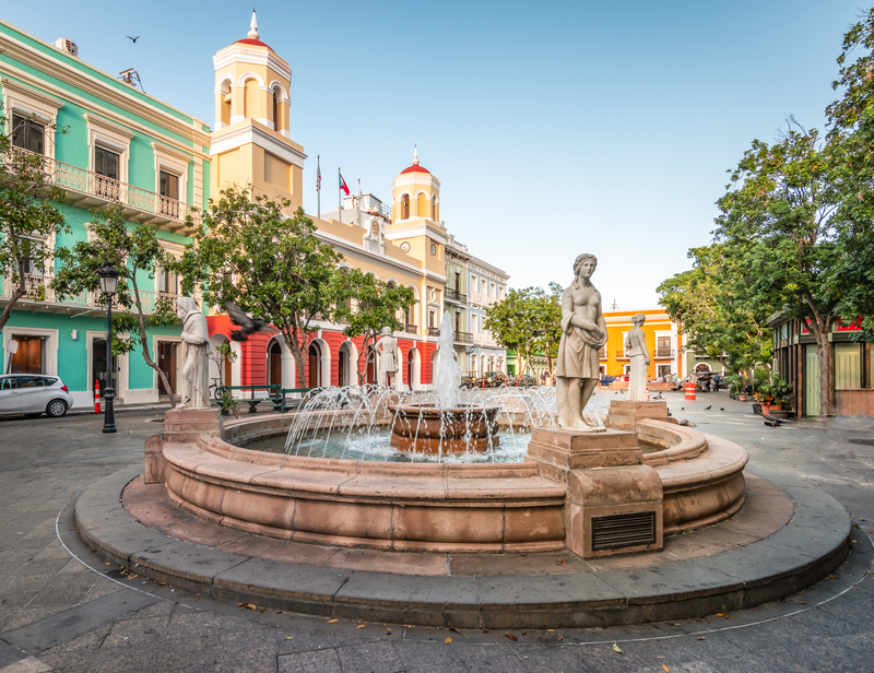 Image of round fountain in plaza with colorful buildings behind it.